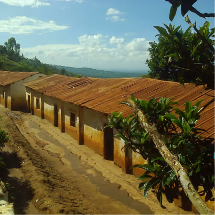 A row of rustic buildings with rusted corrugated metal roofs beside a dirt path, surrounded by greenery and a partly cloudy sky.