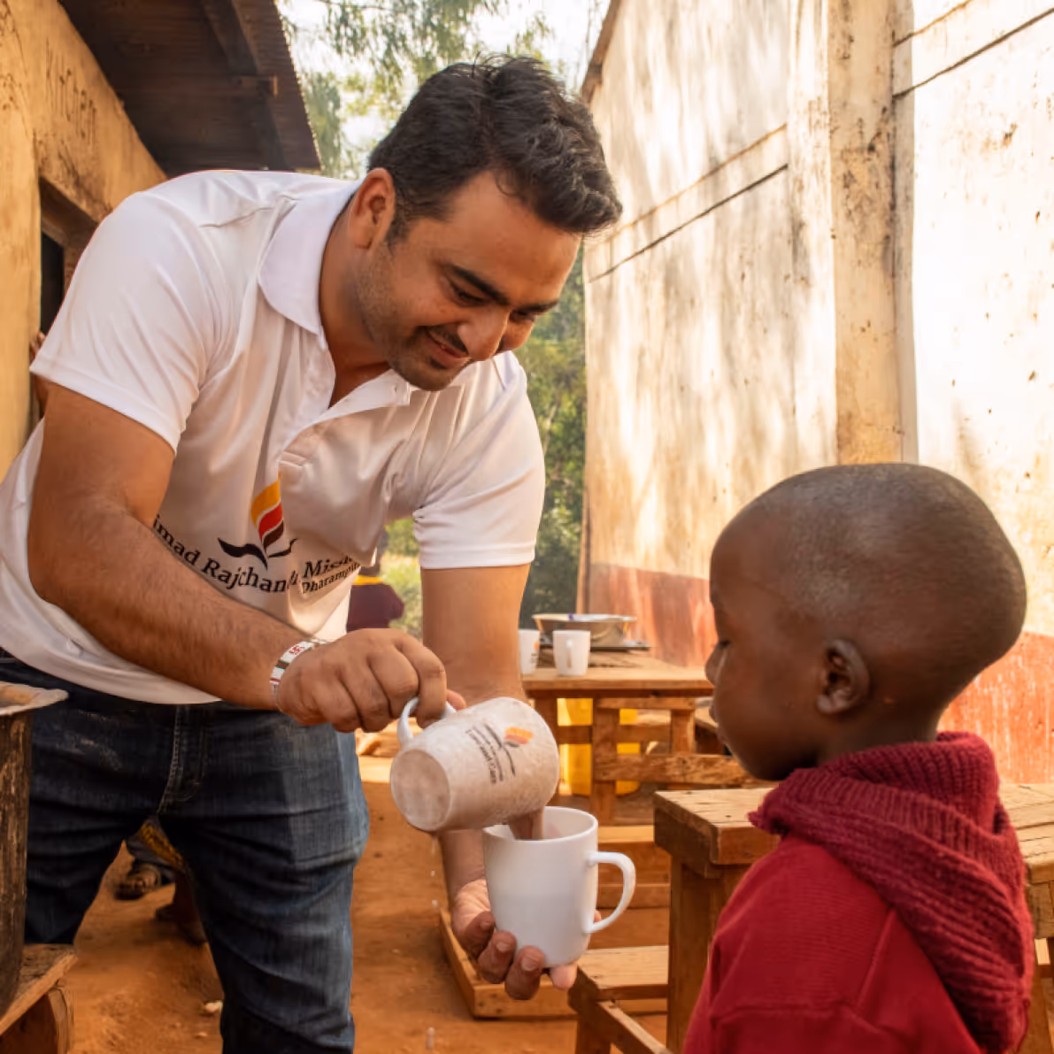 Man in white shirt pouring a drink into a white mug held by a young child wearing a red sweater in an outdoor setting with wooden benches.