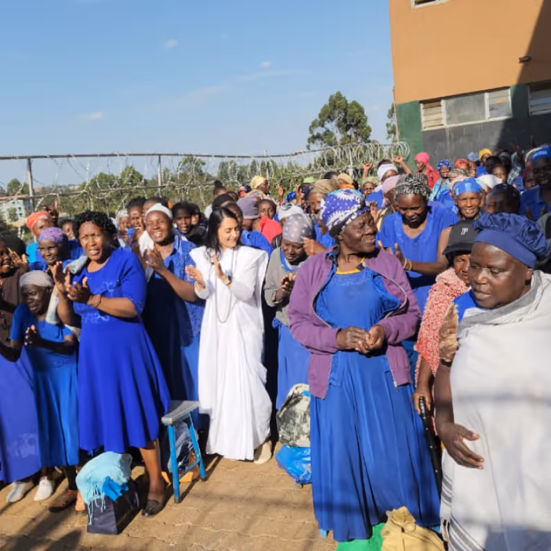 Group of people, mostly women wearing blue dresses and headscarves, gathered outdoors clapping and smiling.