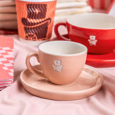 Pink ceramic coffee cup with matching saucer and a small cart logo on the cup, surrounded by a red cup and a patterned takeaway cup in the background.