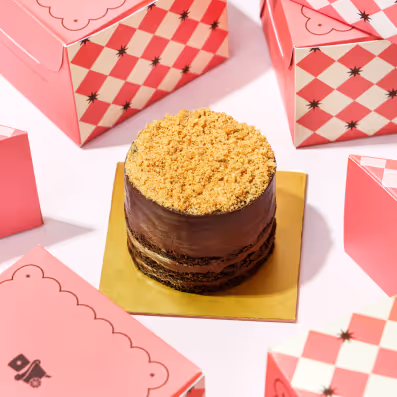 Small round chocolate cake with crumb topping on a gold square base surrounded by pink patterned dessert boxes.