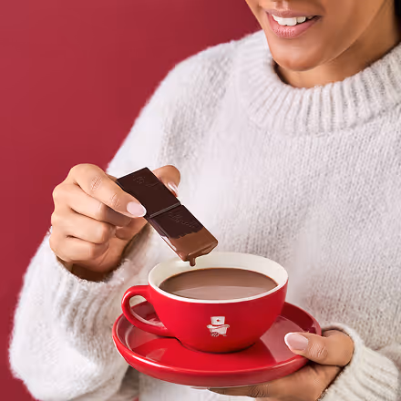 Person holding a red cup and saucer filled with hot chocolate while dipping a piece of chocolate.