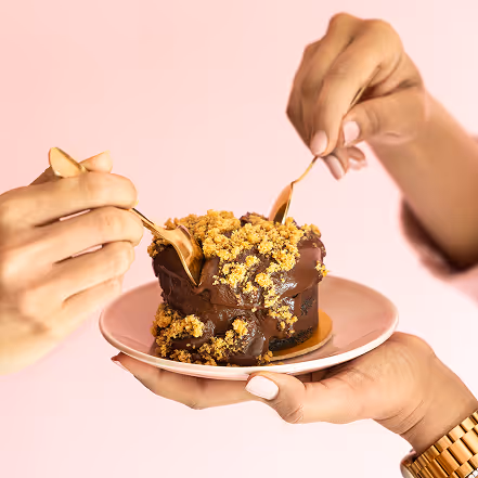 Two hands using small golden spoons to share a chocolate cake with crumb topping on a pink plate.