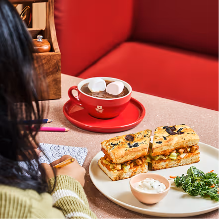Person writing with a pencil at a table with a plate of sandwiches, a small bowl of sauce, and a red cup of hot chocolate with marshmallows.