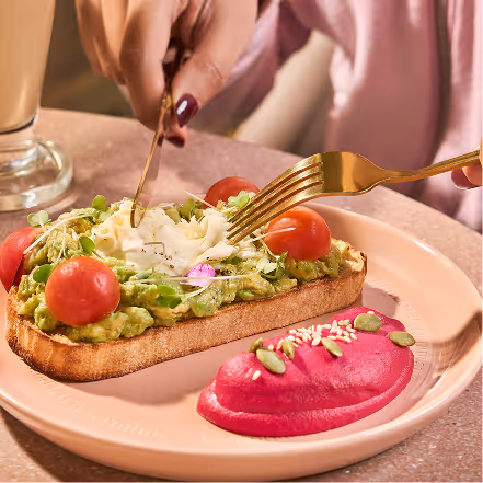 Person using a fork and knife to eat avocado toast topped with cherry tomatoes and a flower garnish, served with a side of beet hummus on a pink plate.
