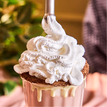 A milkshake topped with whipped cream being dispensed from a nozzle into a glass rimmed with chocolate crumbs and white drizzle.