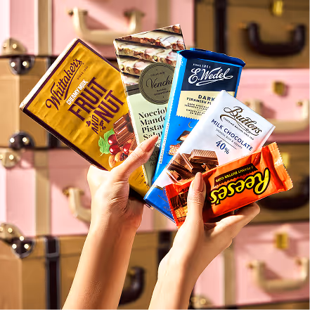 Two hands holding five different chocolate bars including Whitaker's, Venchi, E. Wedel, Butlers, and Reese's, with a background of pink and brown vintage suitcases.