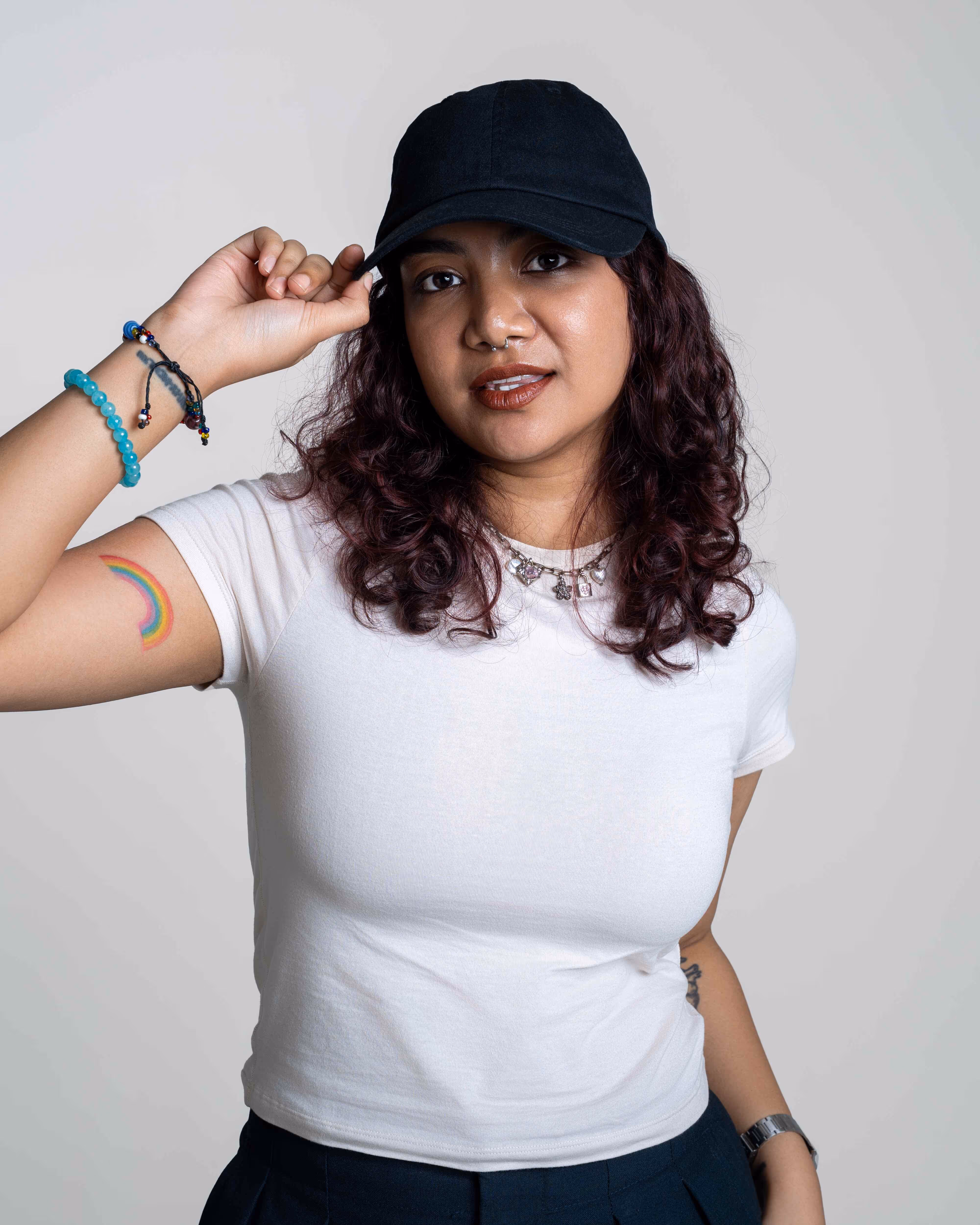 Woman with curly hair wearing a black cap and white t-shirt, showing a rainbow tattoo on her left upper arm.