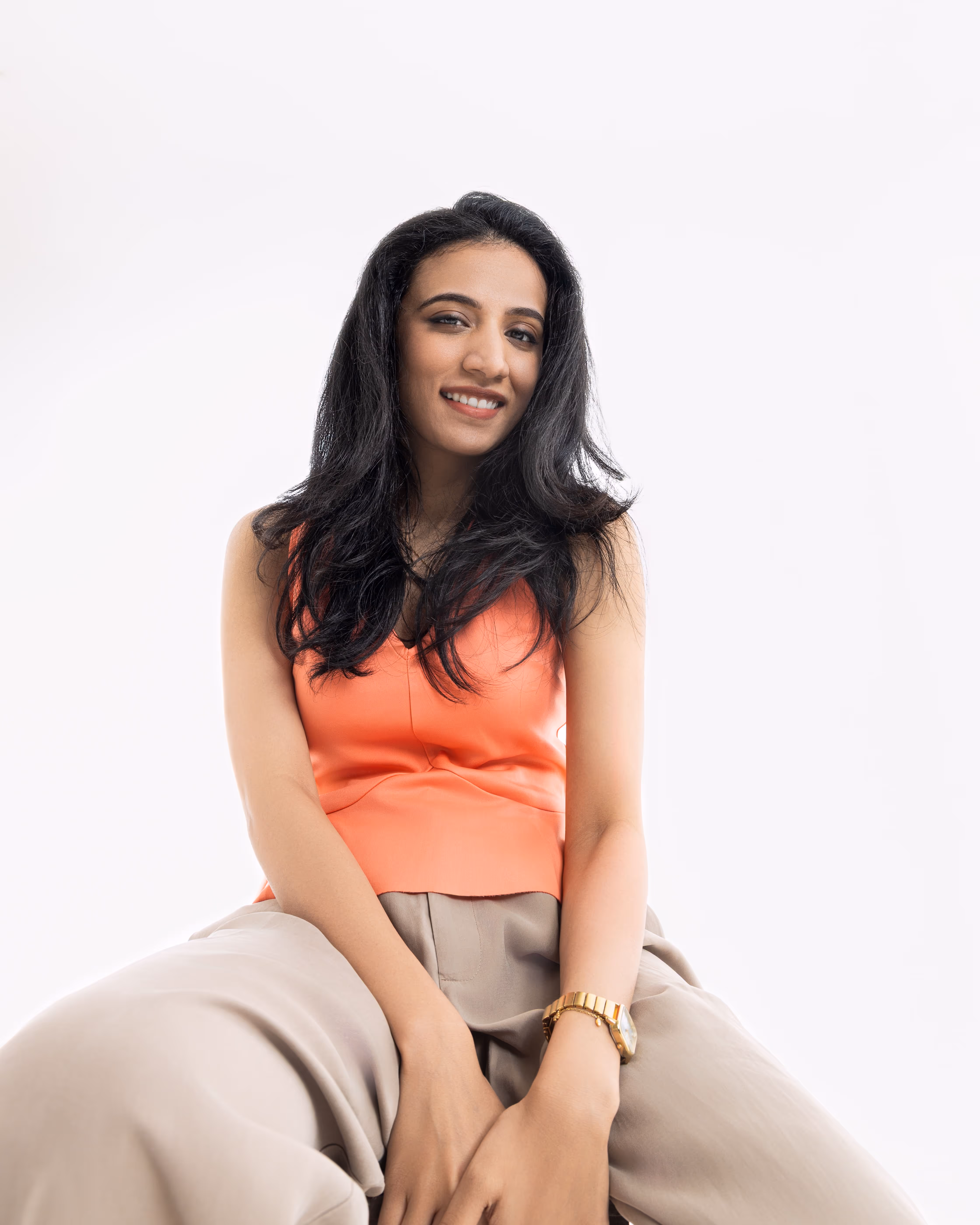 Smiling woman with long black hair wearing an orange sleeveless top and beige pants against a white background.