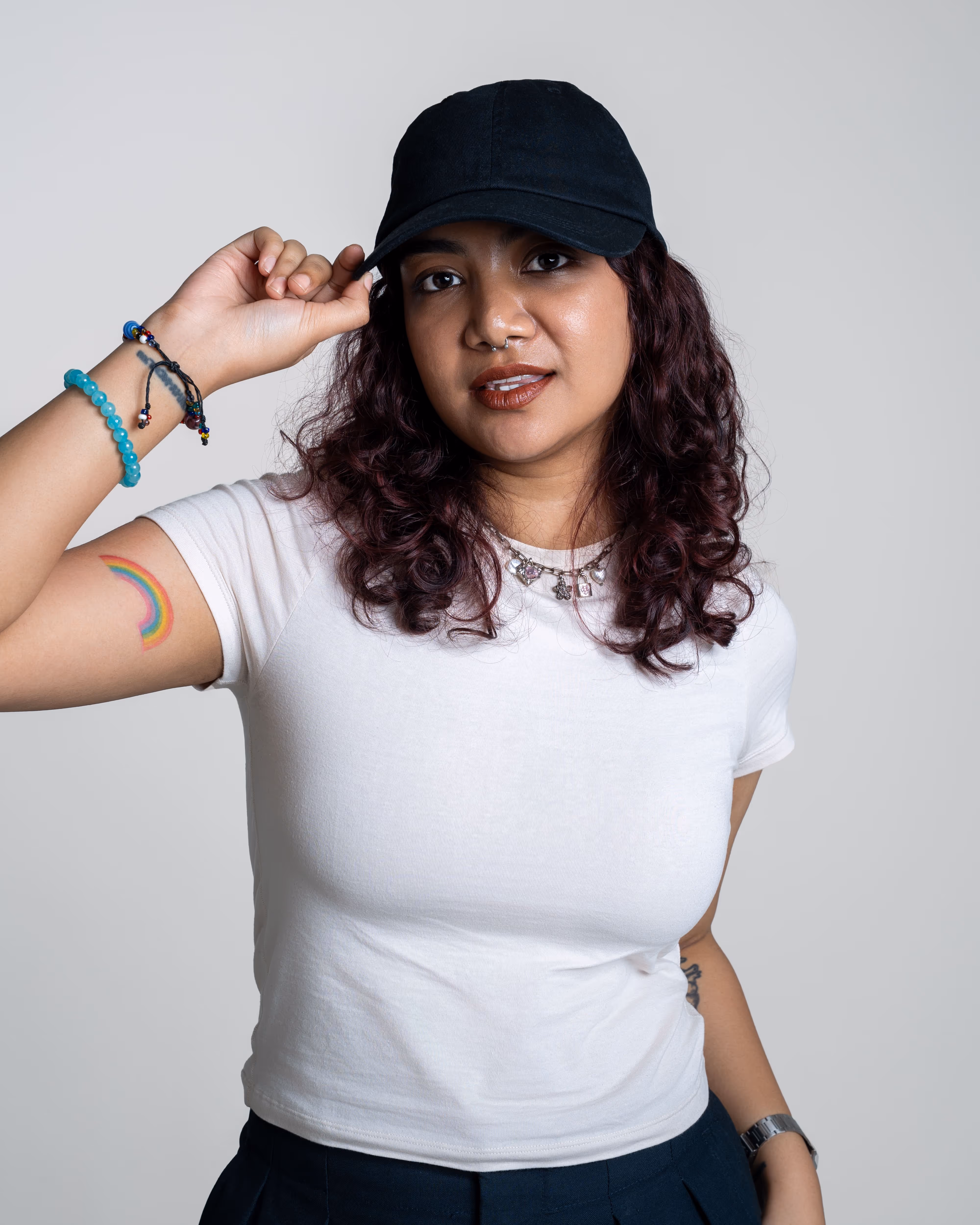 Person with curly dark hair wearing a black cap, white t-shirt, and rainbow tattoo on their arm.