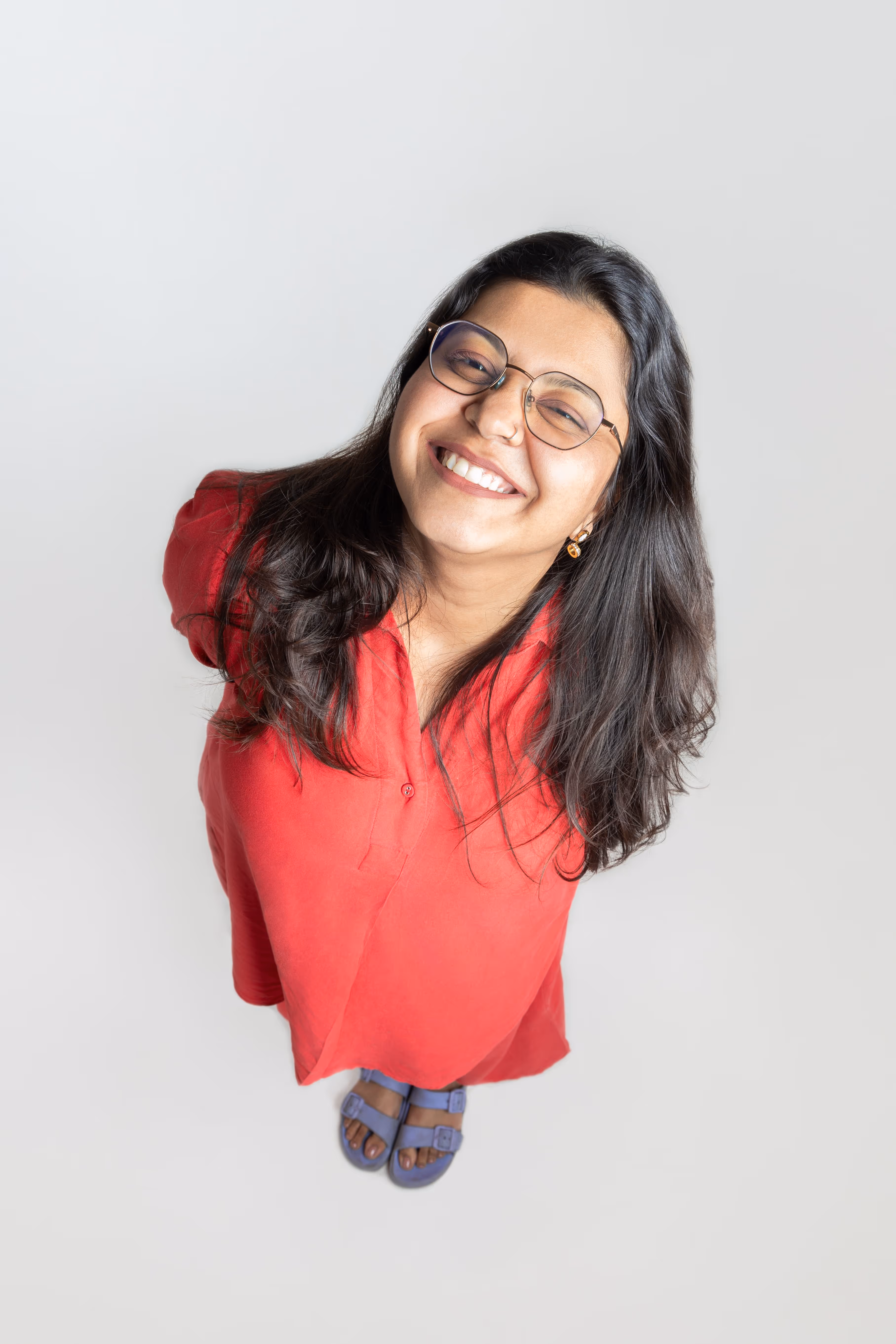 Smiling woman with glasses wearing a red dress and purple sandals standing on a light background, looking up.