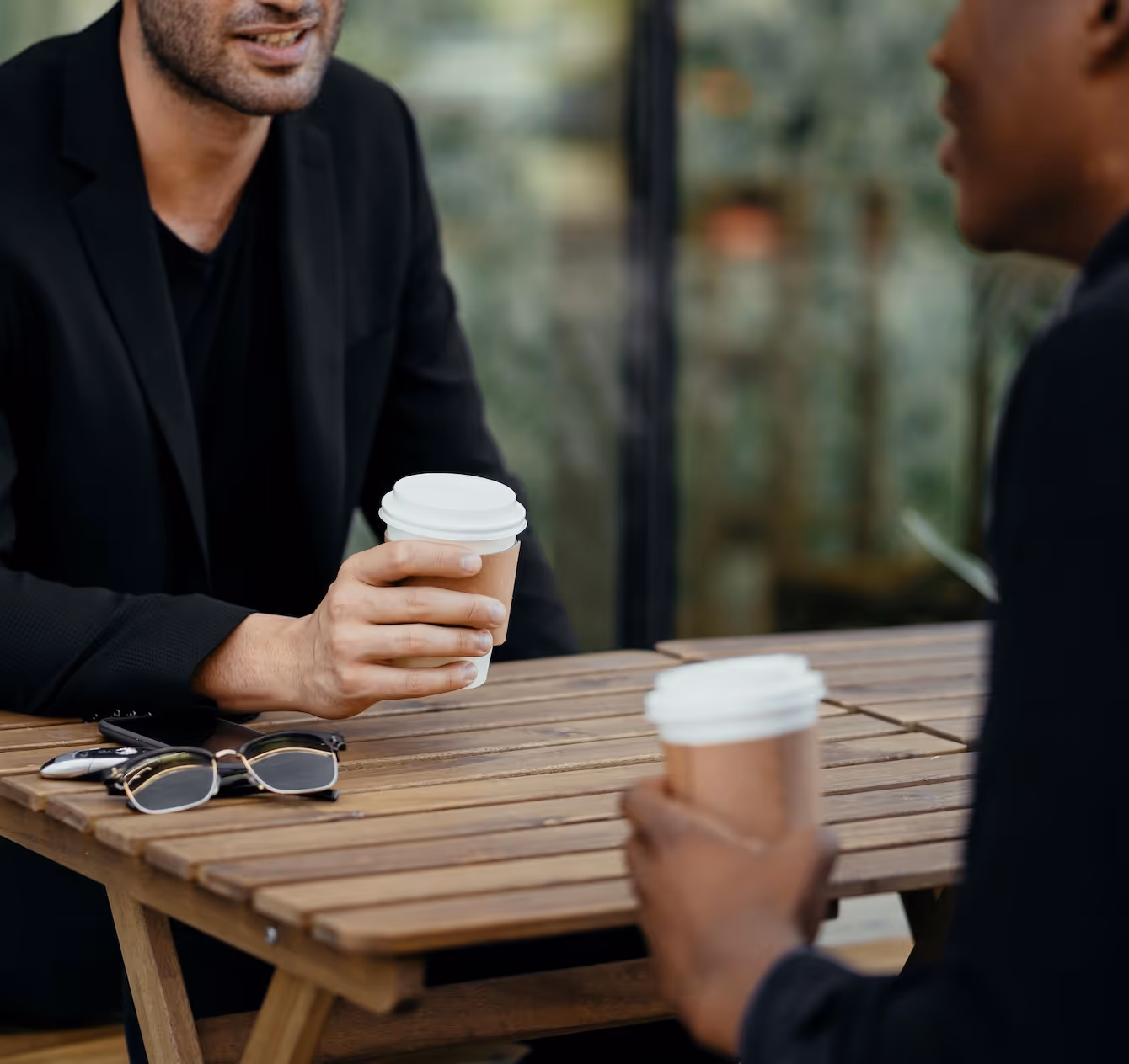 Two men sitting at a wooden table, each holding a coffee cup, with glasses and keys on the table.