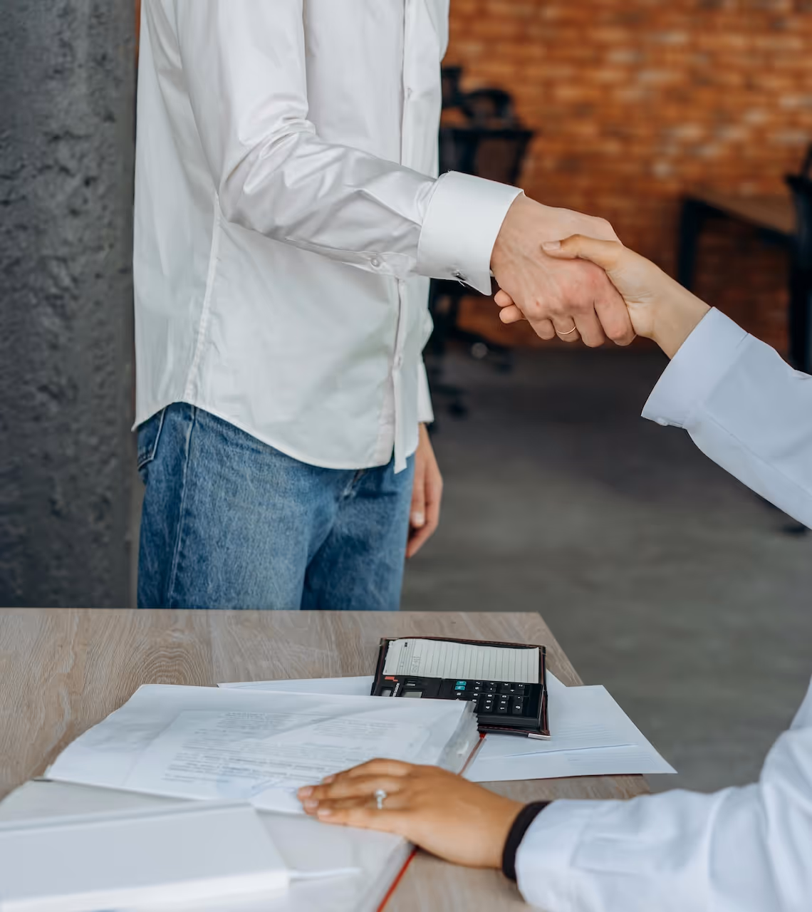 Two people shaking hands over a table with documents and a calculator.