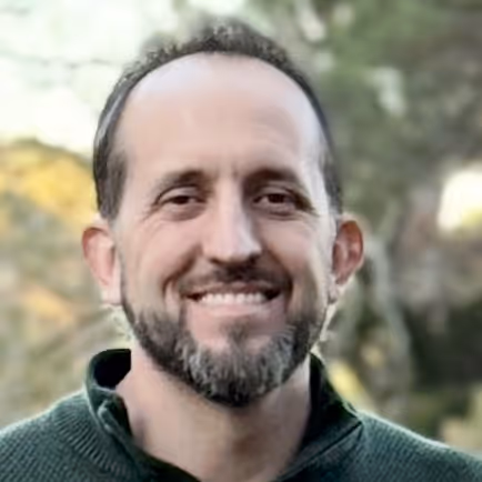 Smiling man with short dark hair and beard wearing a green sweater outdoors.