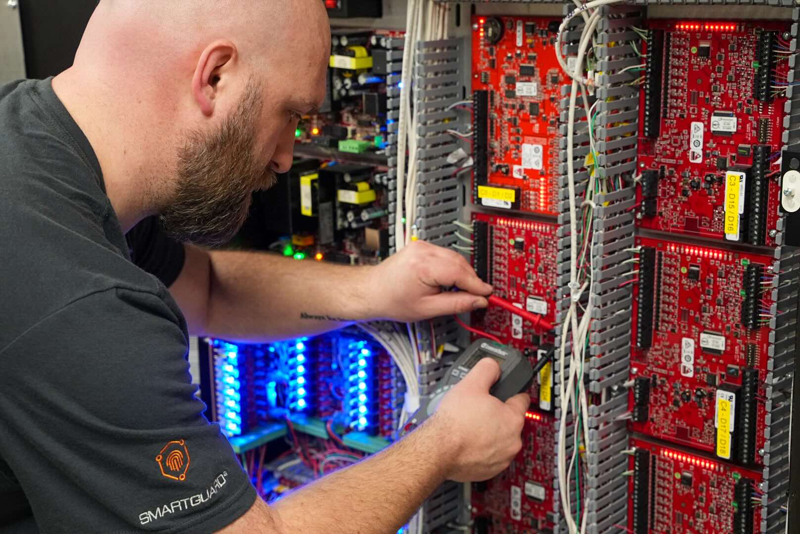 Technician uses a multimeter to test wiring inside an electronic control panel with red circuit boards.