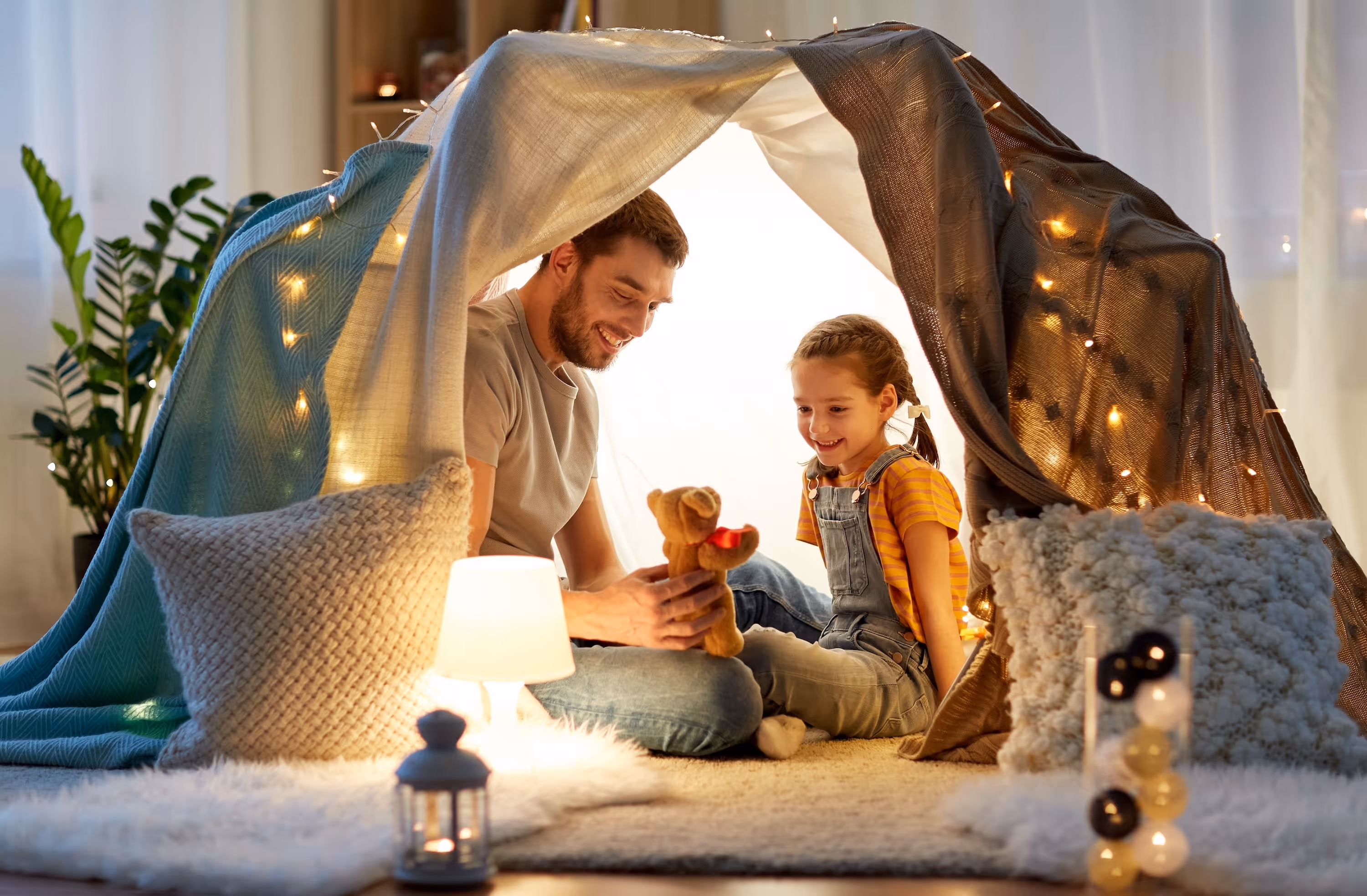 Two residents smiling inside a cozy indoor blanket fort decorated with warm fairy lights and plush pillows at Rancocas Pointe.