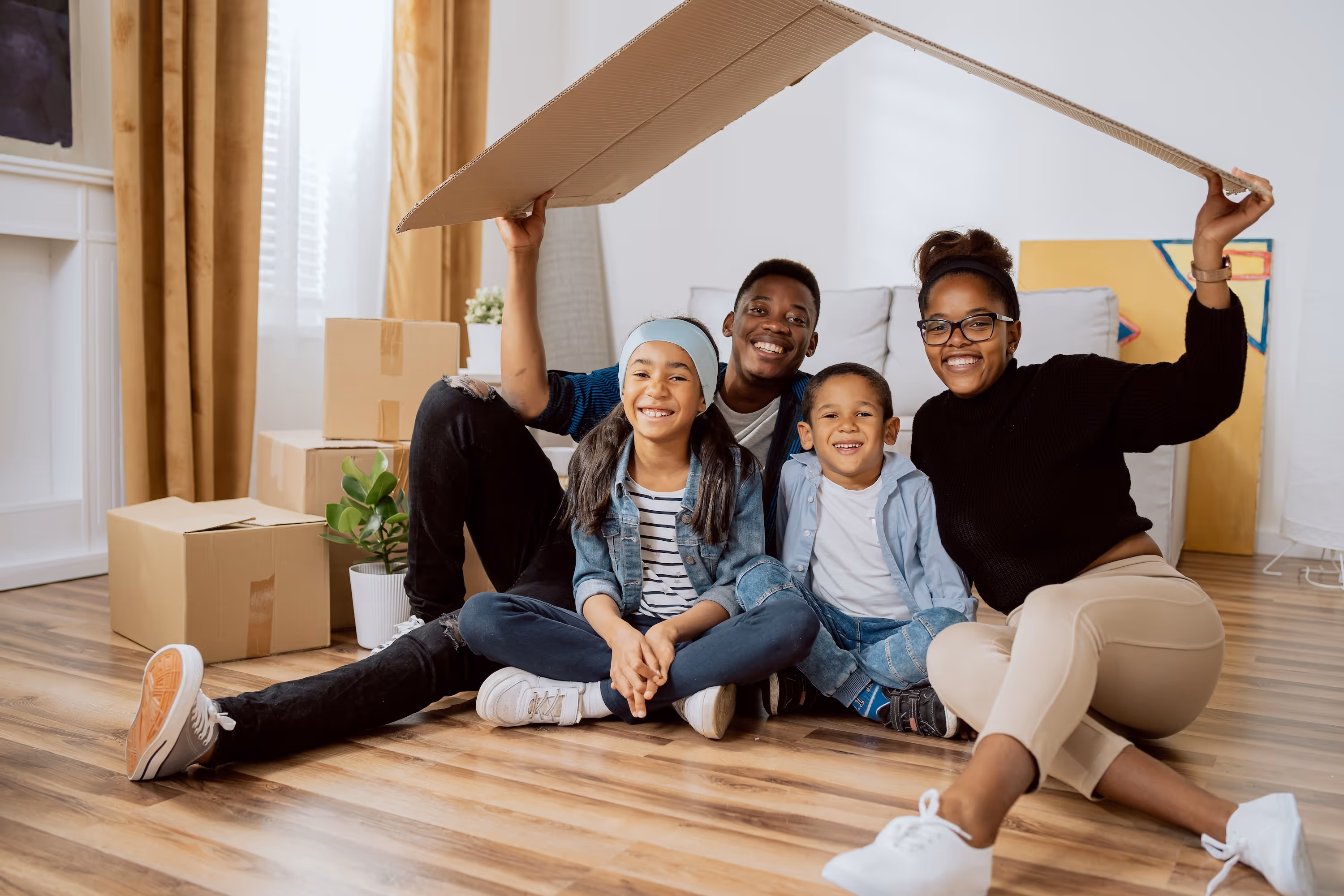 Group of four people sitting on a hardwood floor holding a cardboard "roof" over their heads at their new home at Rancocas Pointe.