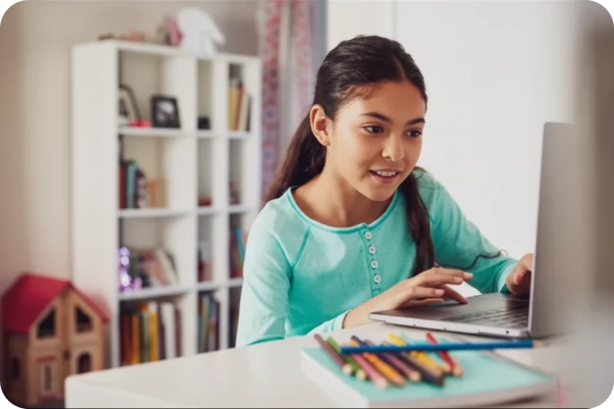 Photograph of a smiling student working on a laptop at a desk with colored pencils.