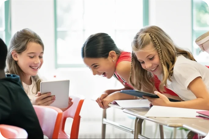Photograph of three students laughing together while sharing a tablet in a classroom.