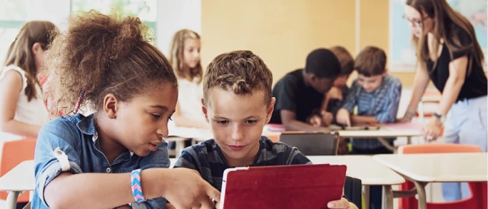 Photograph of two students collaborating and pointing at a tablet screen in a busy classroom.