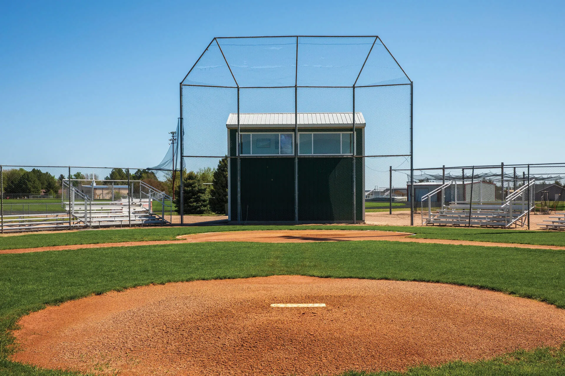 A baseball field on a sunny day, featuring a freshly mowed green outfield, red dirt base paths, and empty bleachers in the distance.