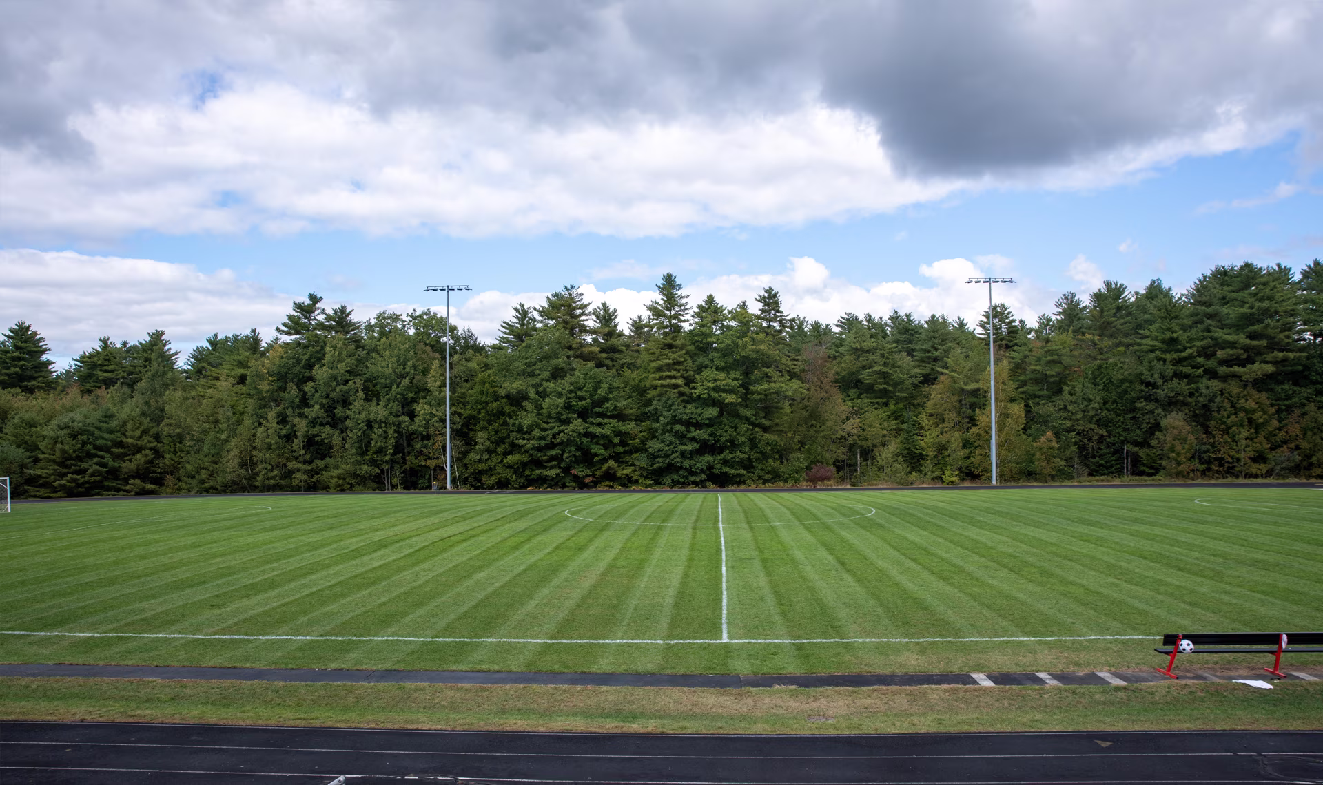 Outdoor soccer field on a sunny day, with a goal net in the foreground and trees in the background.