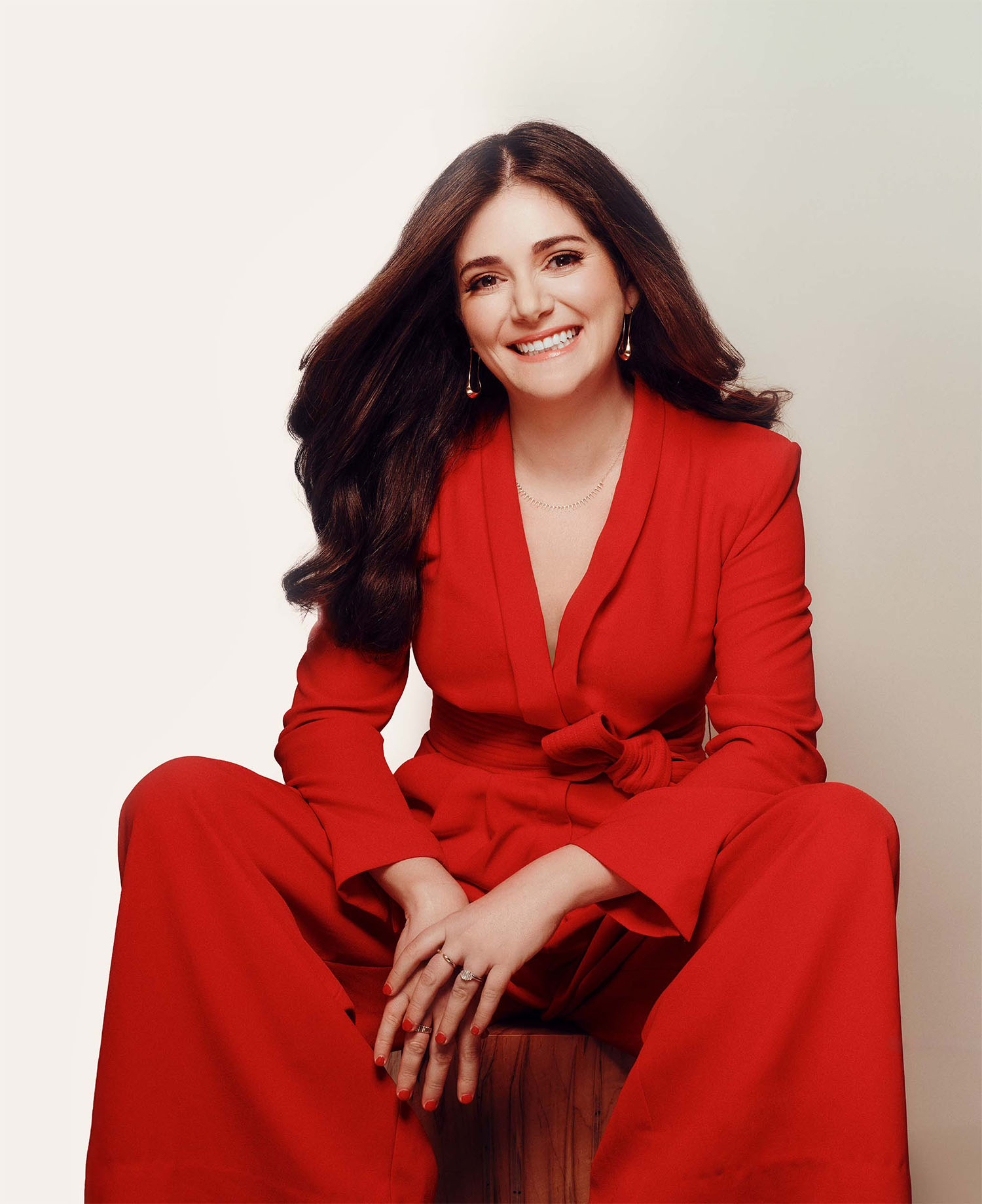 Smiling woman with long dark hair wearing a red suit, seated with hands clasped in front.