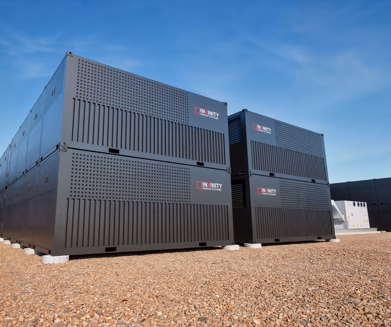 Large black Infinity Energy Systems battery storage units stacked outdoors on gravel under a blue sky.