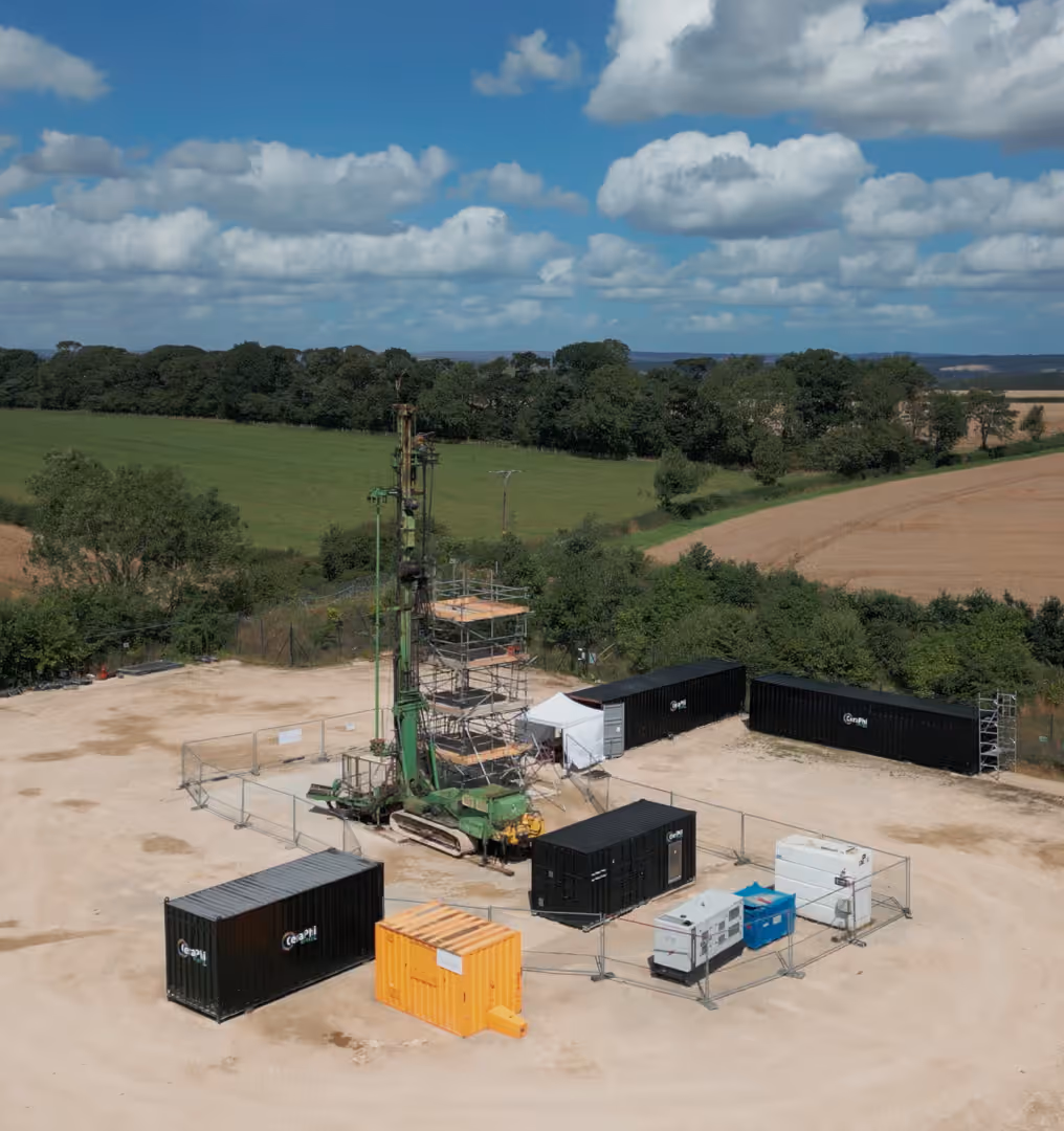 Geothermal drilling setup with a green rig, containers, and equipment on a cleared site near fields and trees.