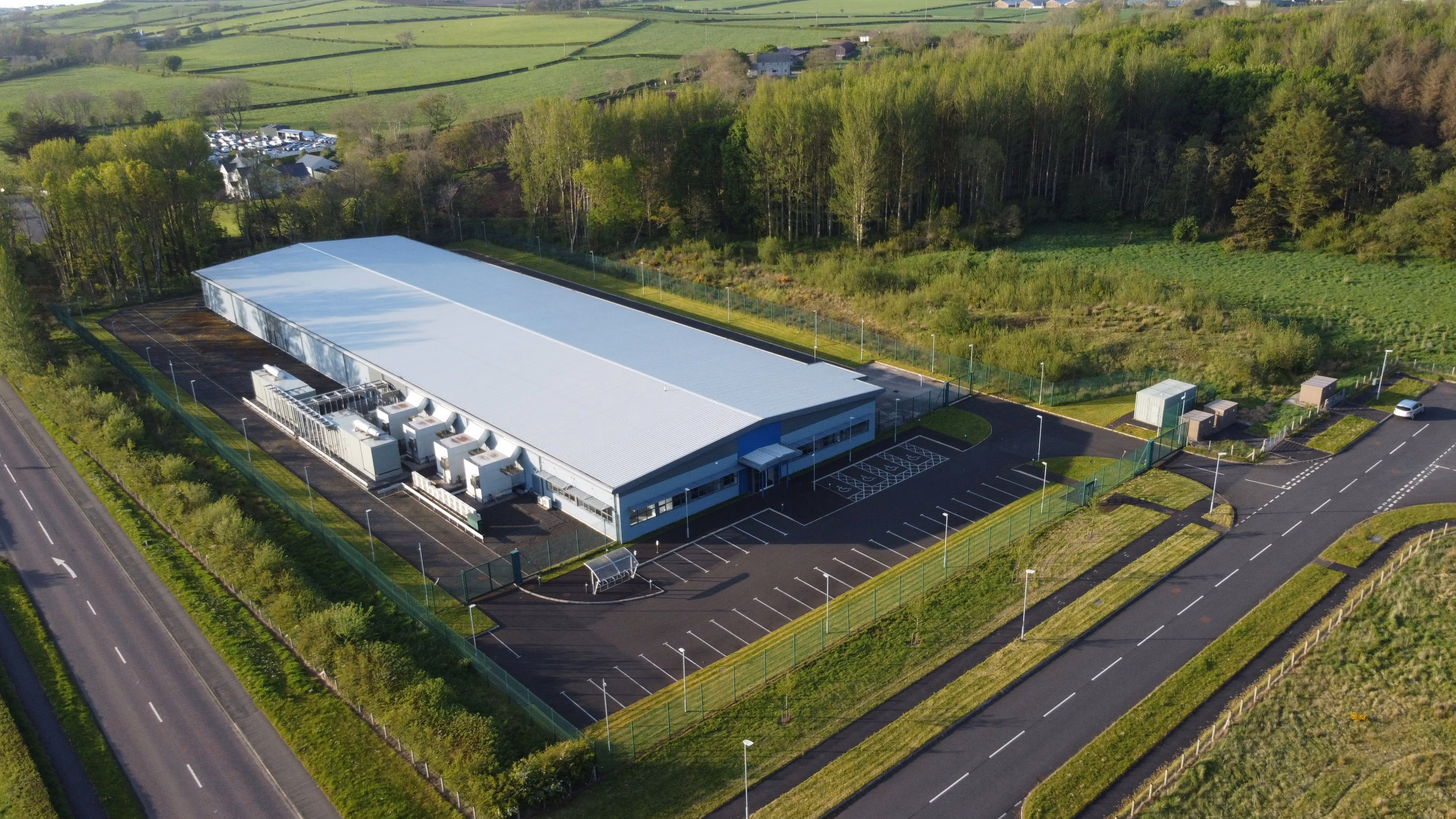 Aerial view of a large industrial building with surrounding parking and greenery in a rural setting.