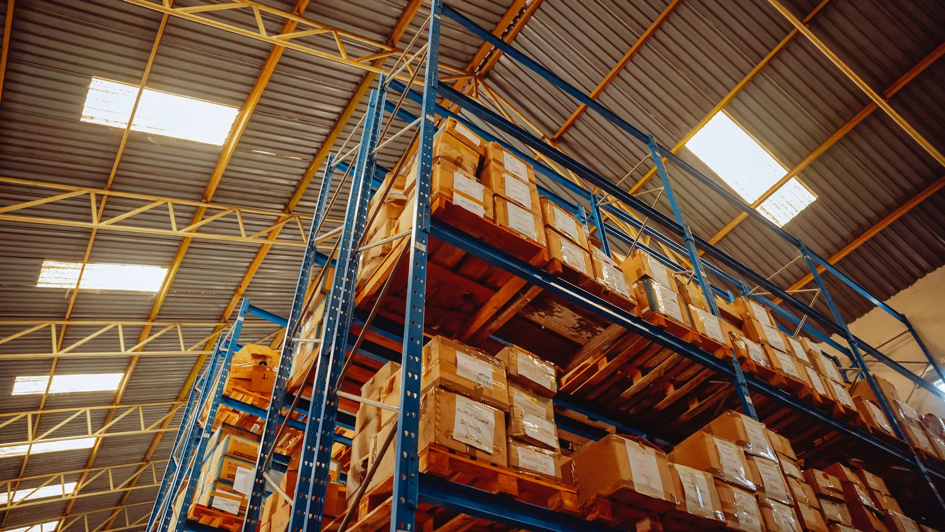 Warehouse shelves with stacked cardboard boxes on wooden pallets under a metal roof.