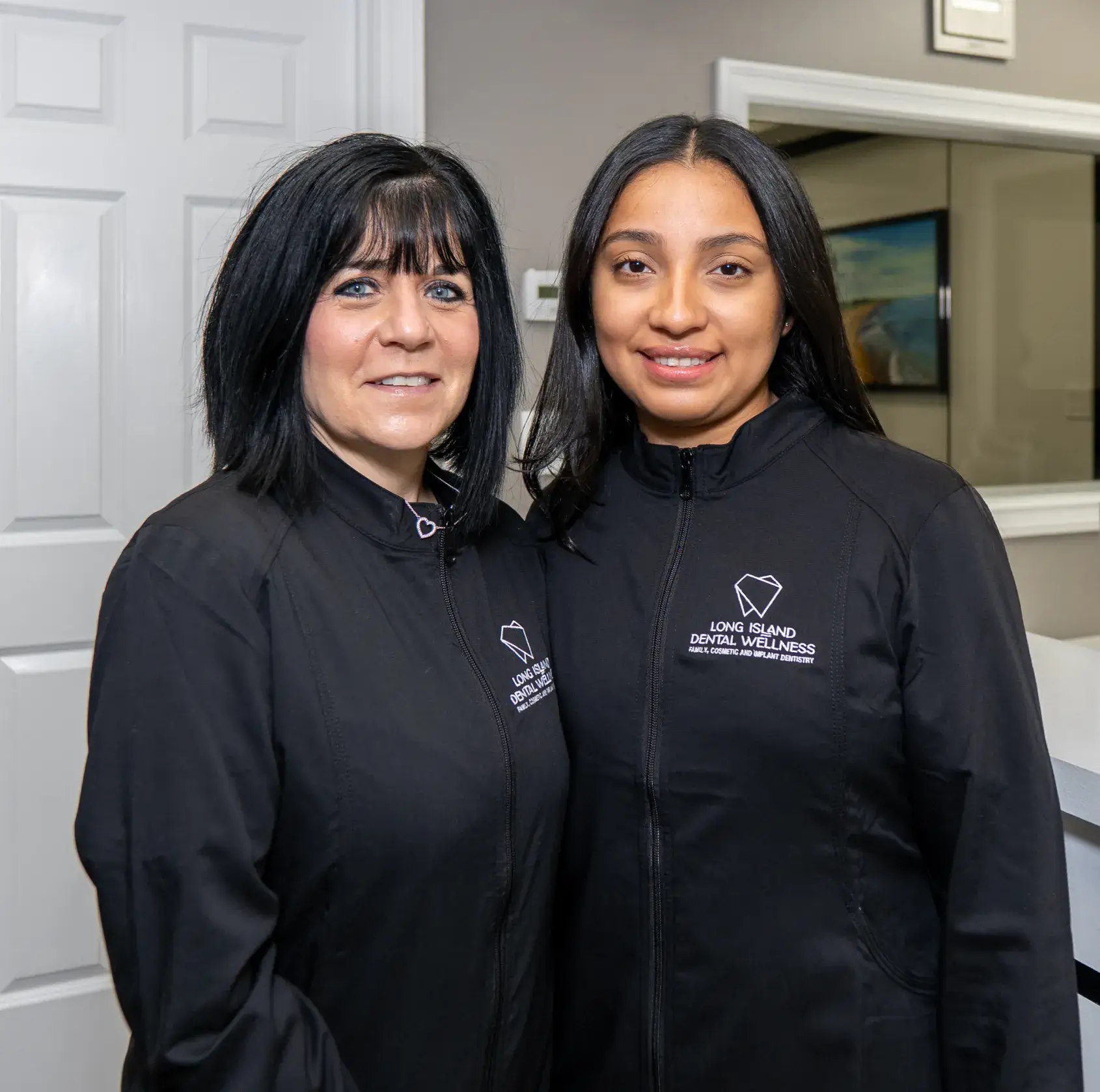 Two women standing side by side, both wearing matching black zip-up sweaters indoors.