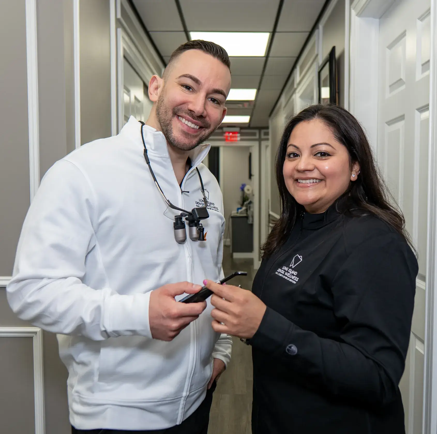 Two smiling dental professionals stand in a hallway, holding dental instruments.