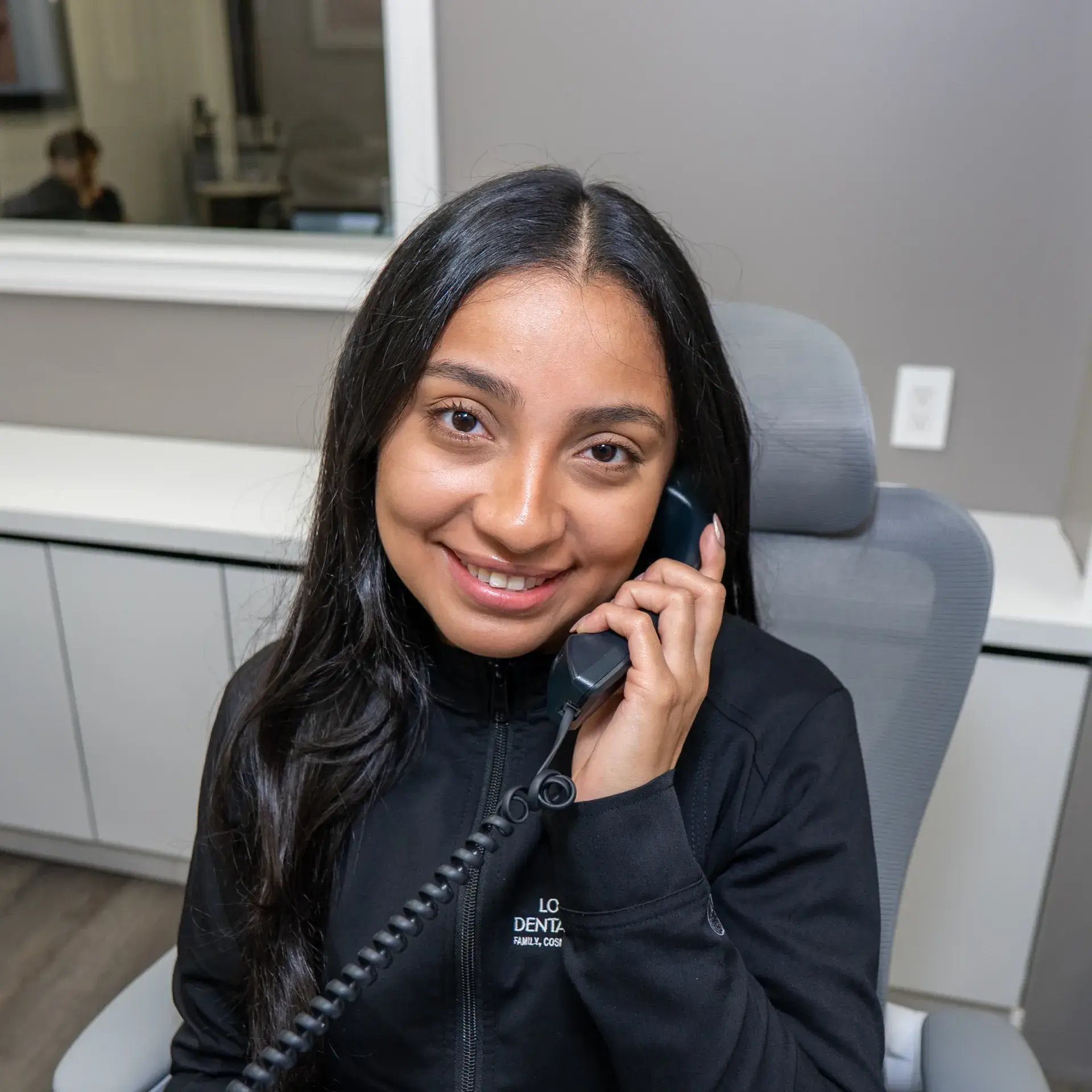 A woman on the phone, smiling, sits at an office desk.