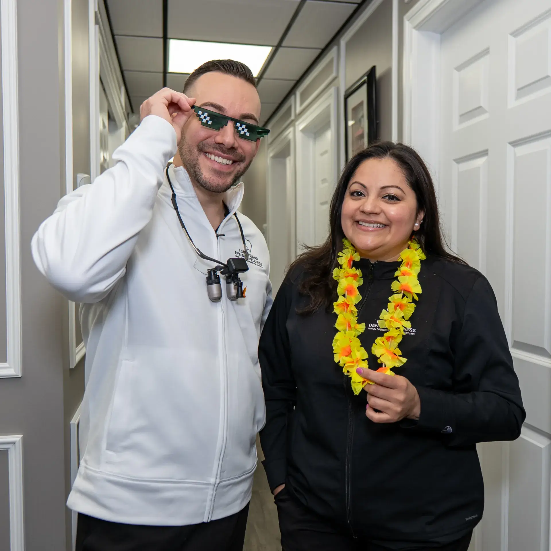 Man wearing dental tools and checkered sunglasses stands next to smiling woman with yellow lei.