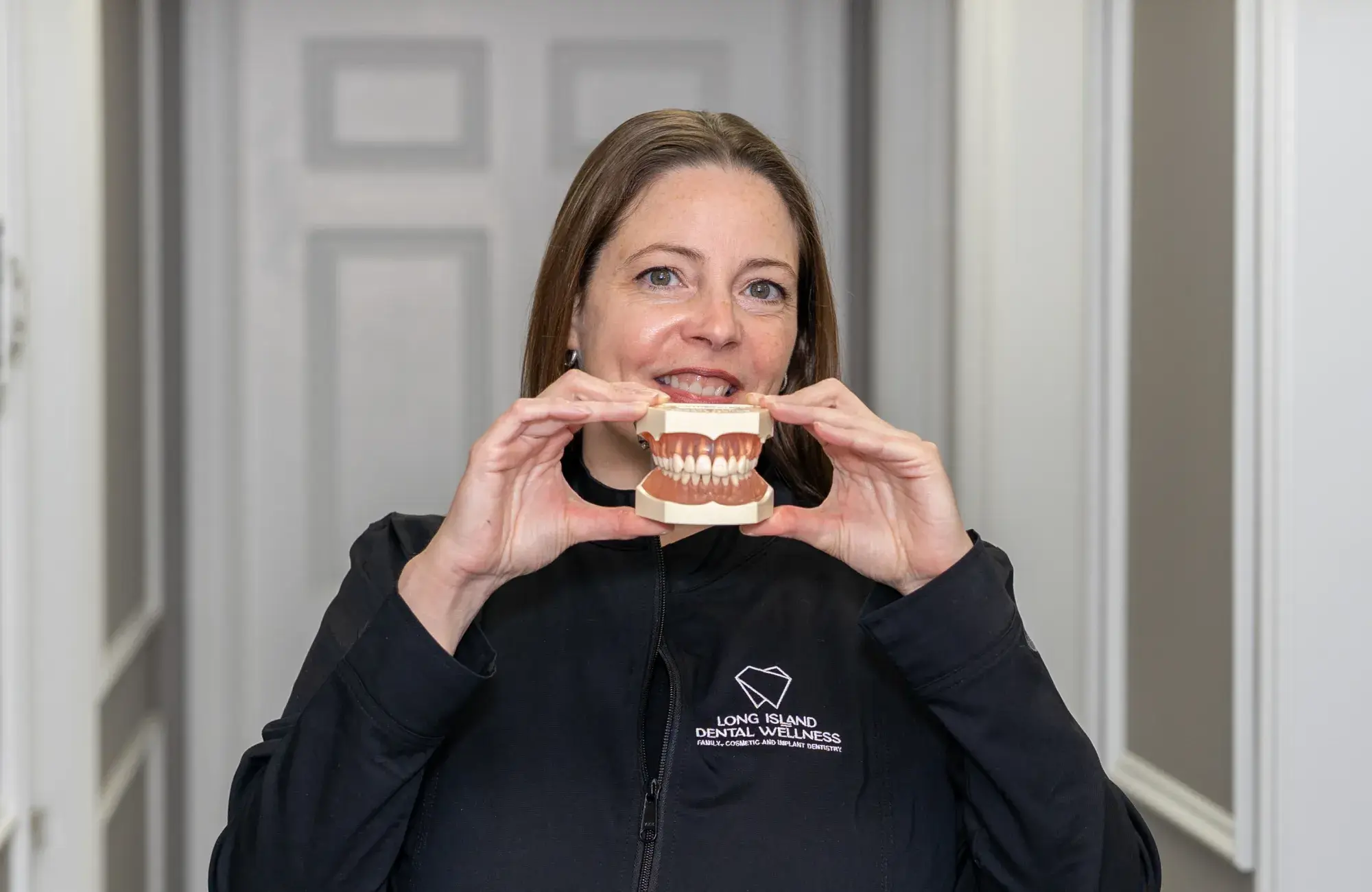 A woman holding a model of human teeth, smiling in a dental office setting.