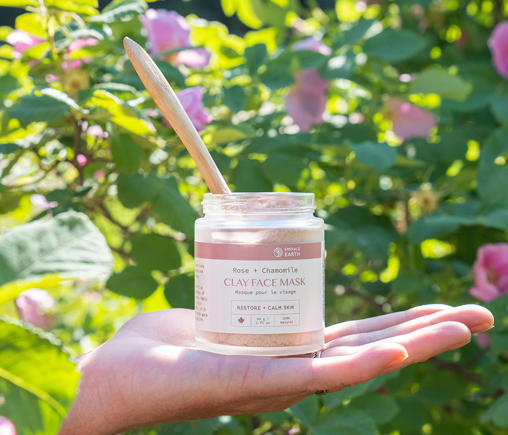 Hand holding a jar of Rose + Chamomile clay face mask with a wooden spatula outdoors.