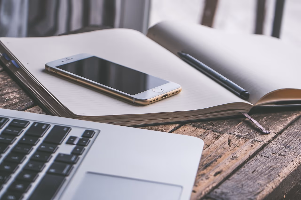 A smartphone rests on a blank notebook beside a laptop, with a pen nearby, on a rustic wooden table.