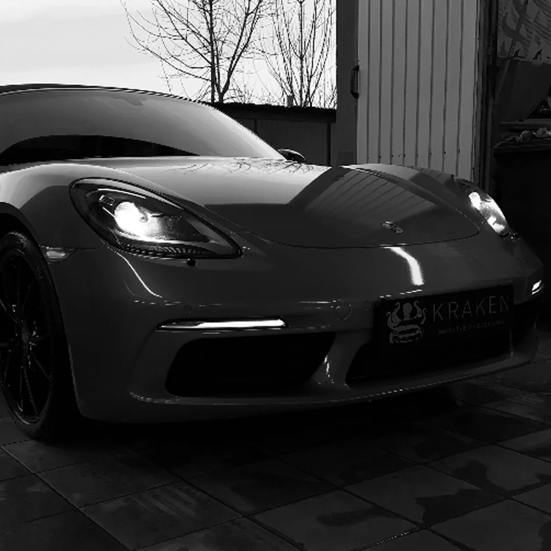 Спереду спортивний автомобіль Porsche з увімкненими фарами на парковці біля будівлі.