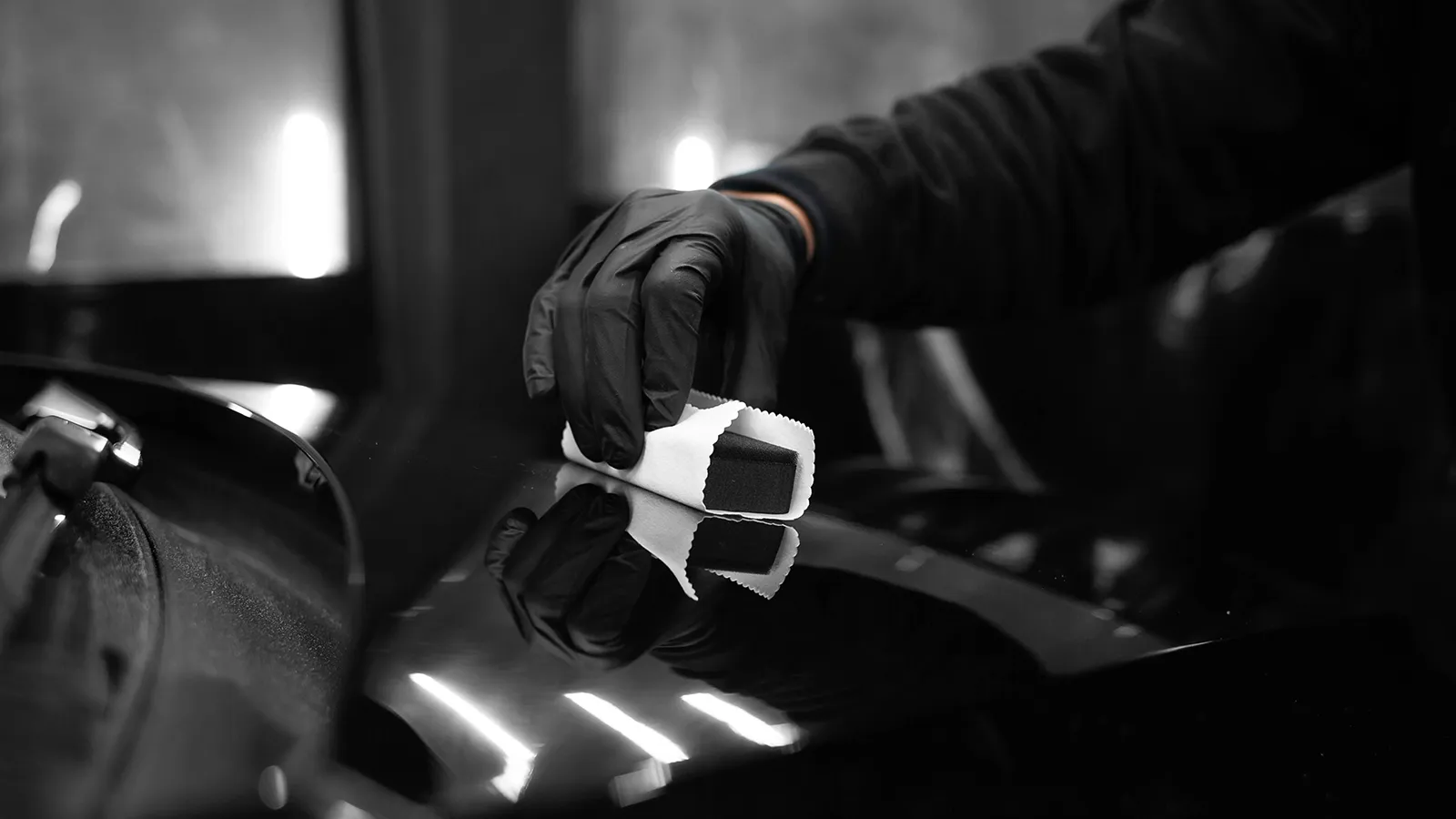 Рука в чорній рукавичці полірує блискучу чорну поверхню автомобіля губкою в білому чохлі.