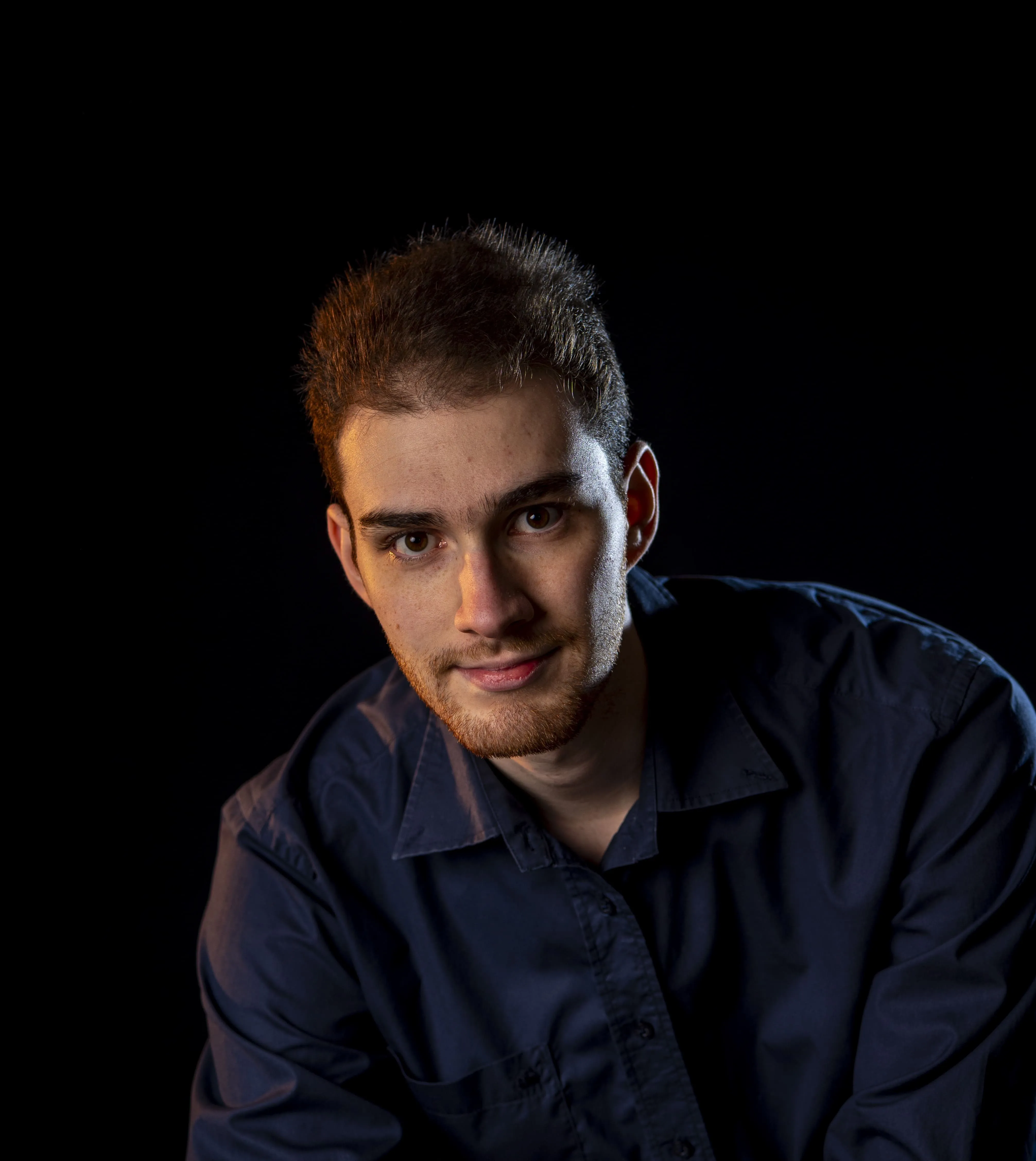 Periklis Kotinis with brown hair and beard wearing a dark blue shirt leaning forward on a chair against black background.