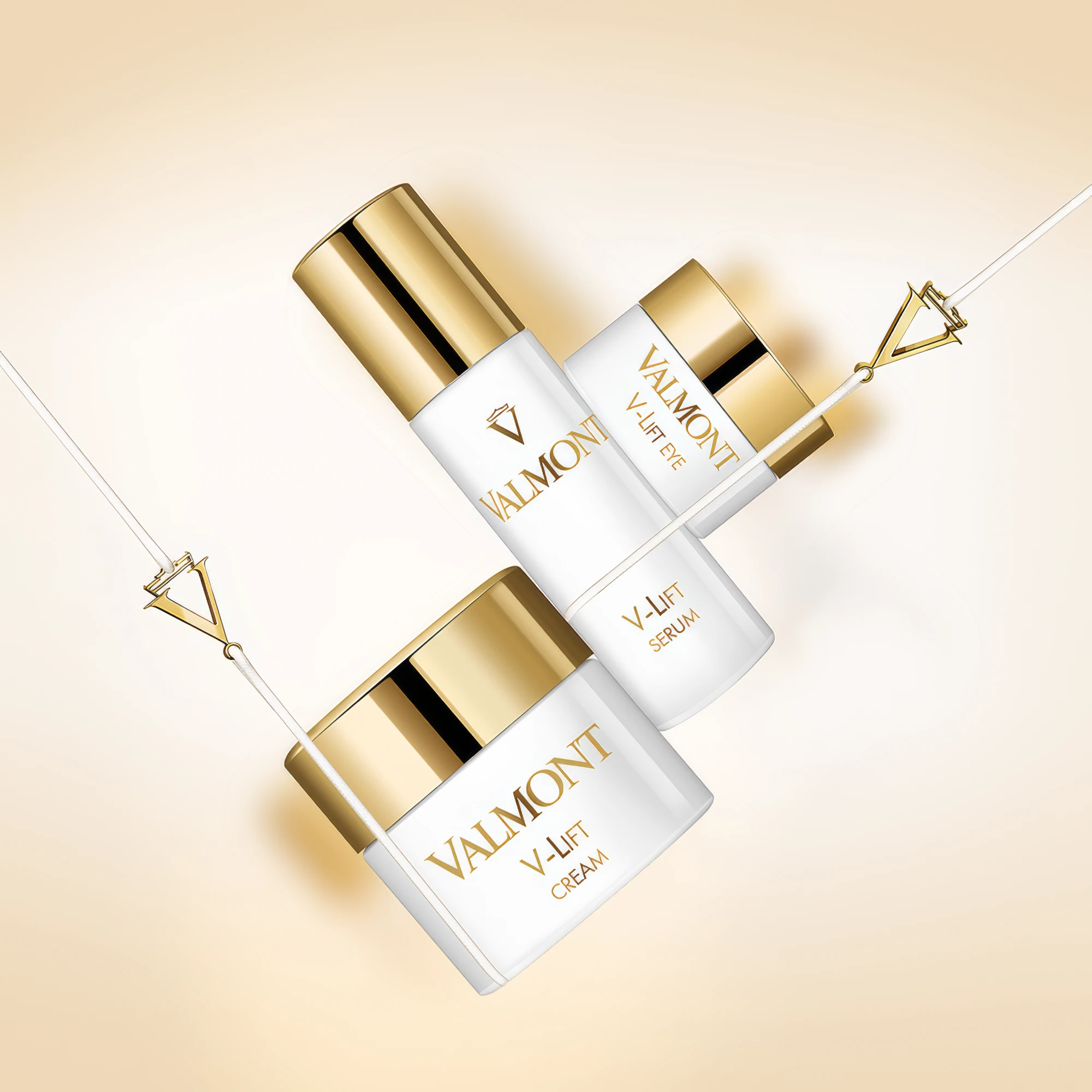 Valmont V-Lift Cream, Serum and Eye suspended on gold V-shaped cords against a warm cream backdrop