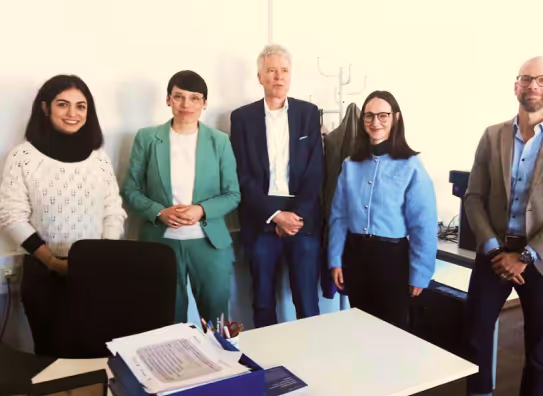 Eine Gruppe von fünf Personen steht in einem Büro-Setting, mit einem Schreibtisch und Papierkram im Hintergrund. Alle sind professionell gekleidet.
