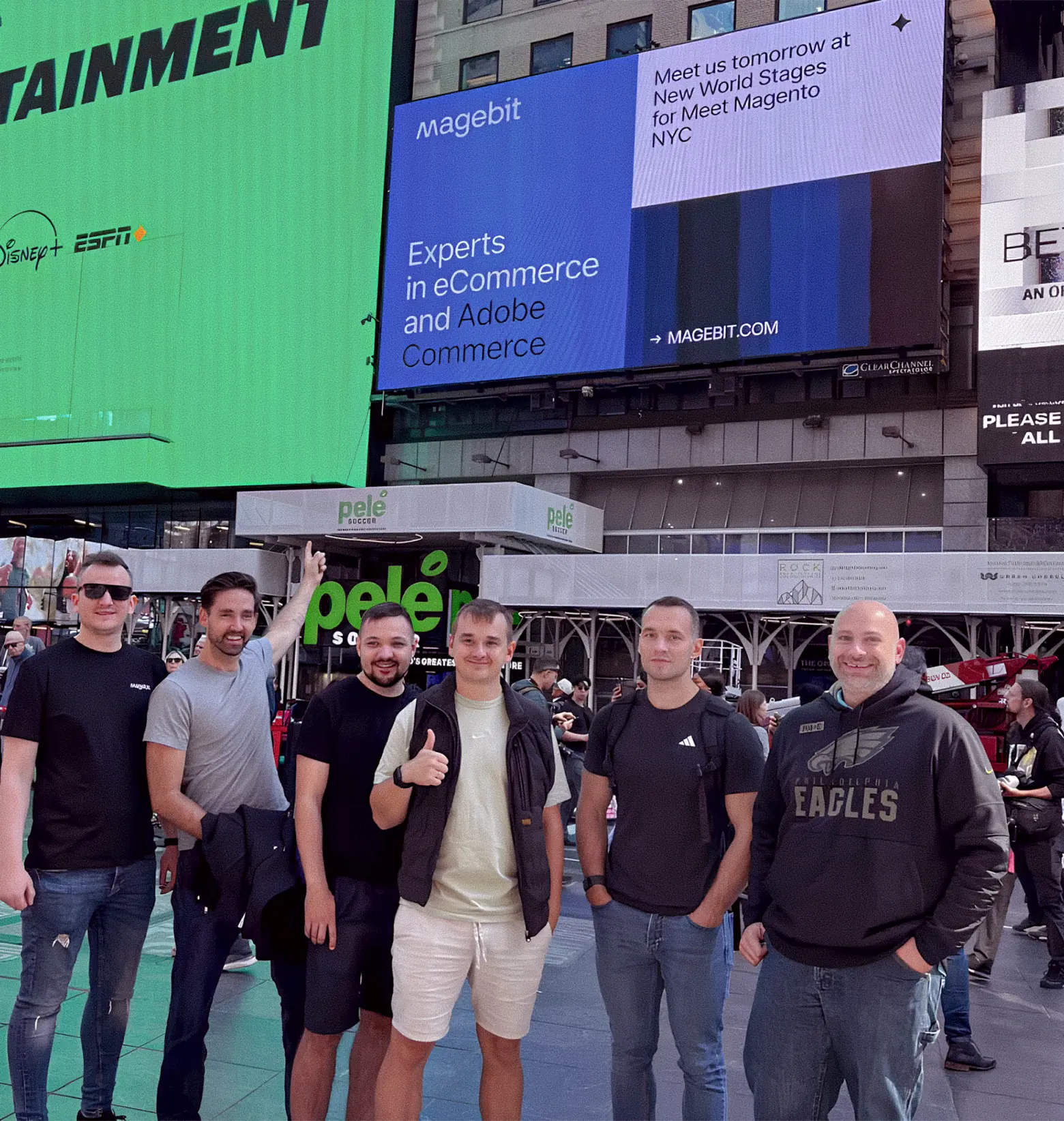 Six men pose outdoors in casual clothes under large digital billboards in a busy urban area.