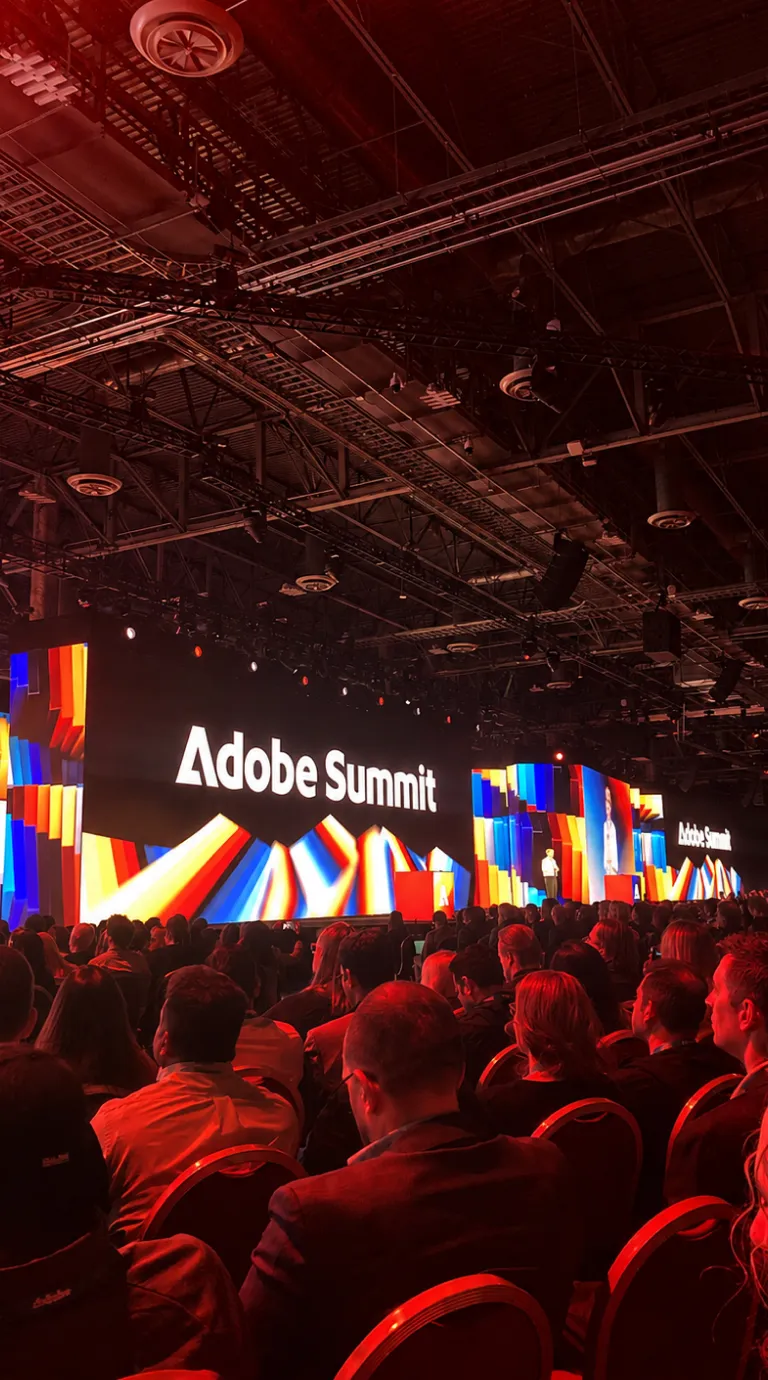 Audience seated at Adobe Summit conference with colorful stage and large screens displaying Adobe Summit logo.