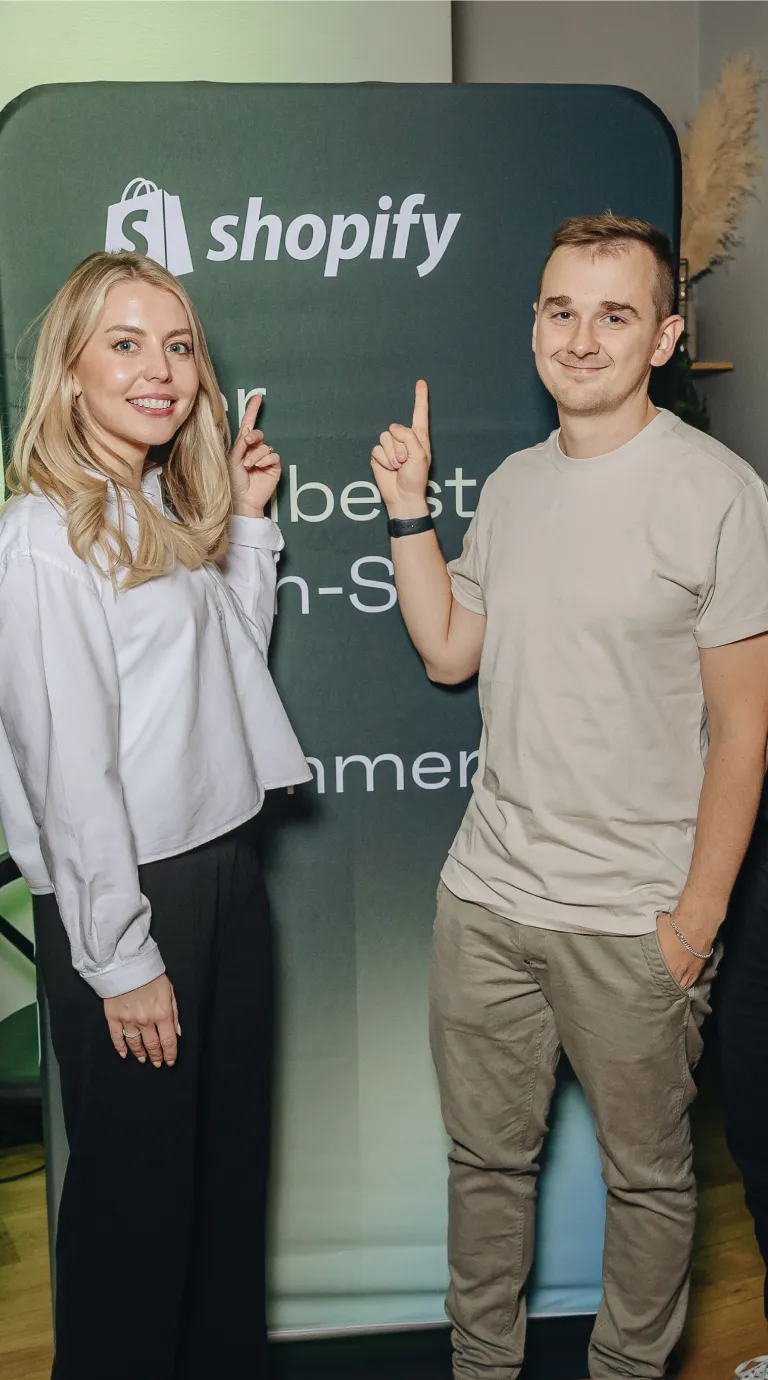 Smiling man and woman pointing upwards to a Shopify sign on a stand behind them indoors.