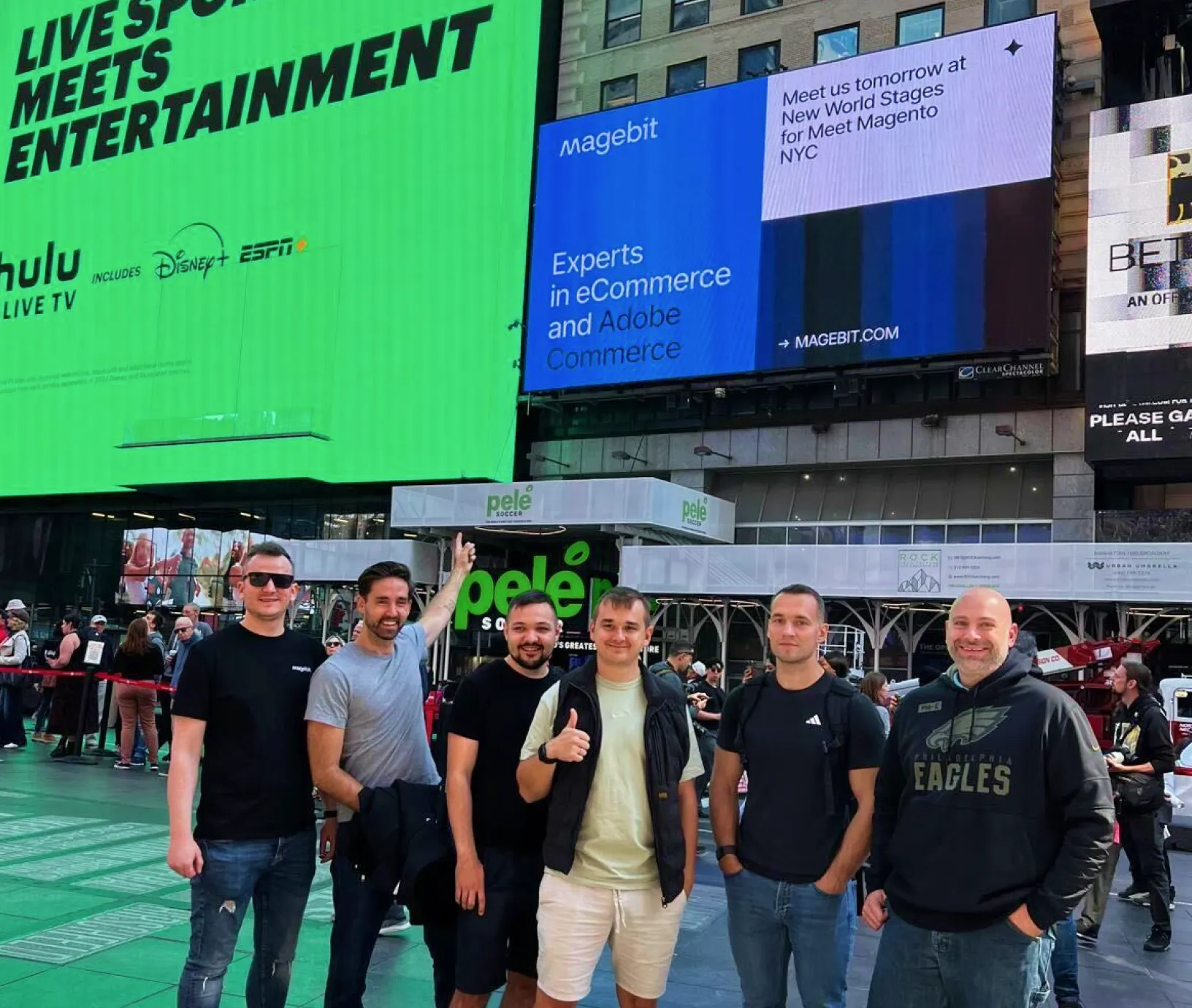 Group of six men posing in front of digital billboards in an urban outdoor area.