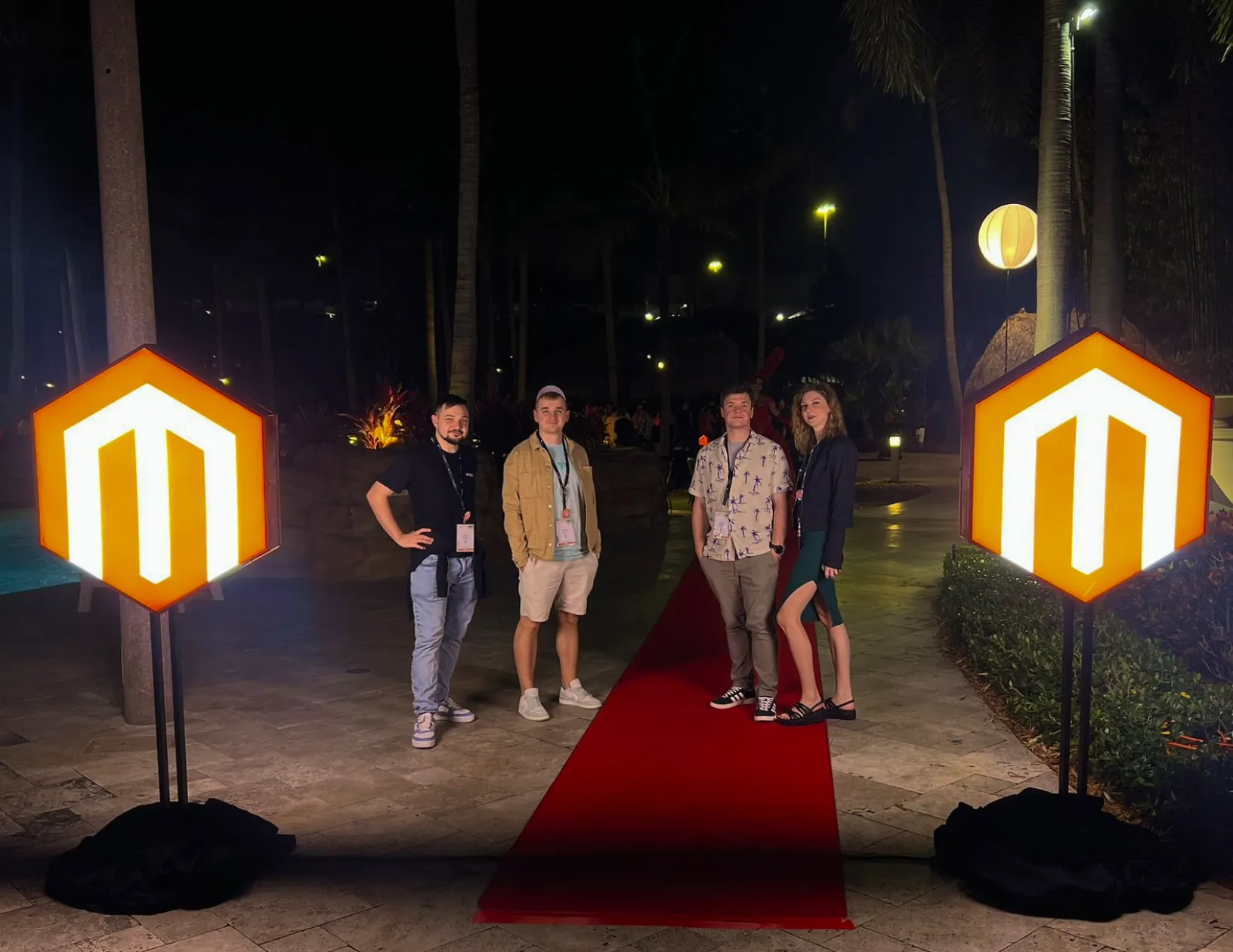 Four people standing on a red carpet between two illuminated Magento logos at night outdoors.