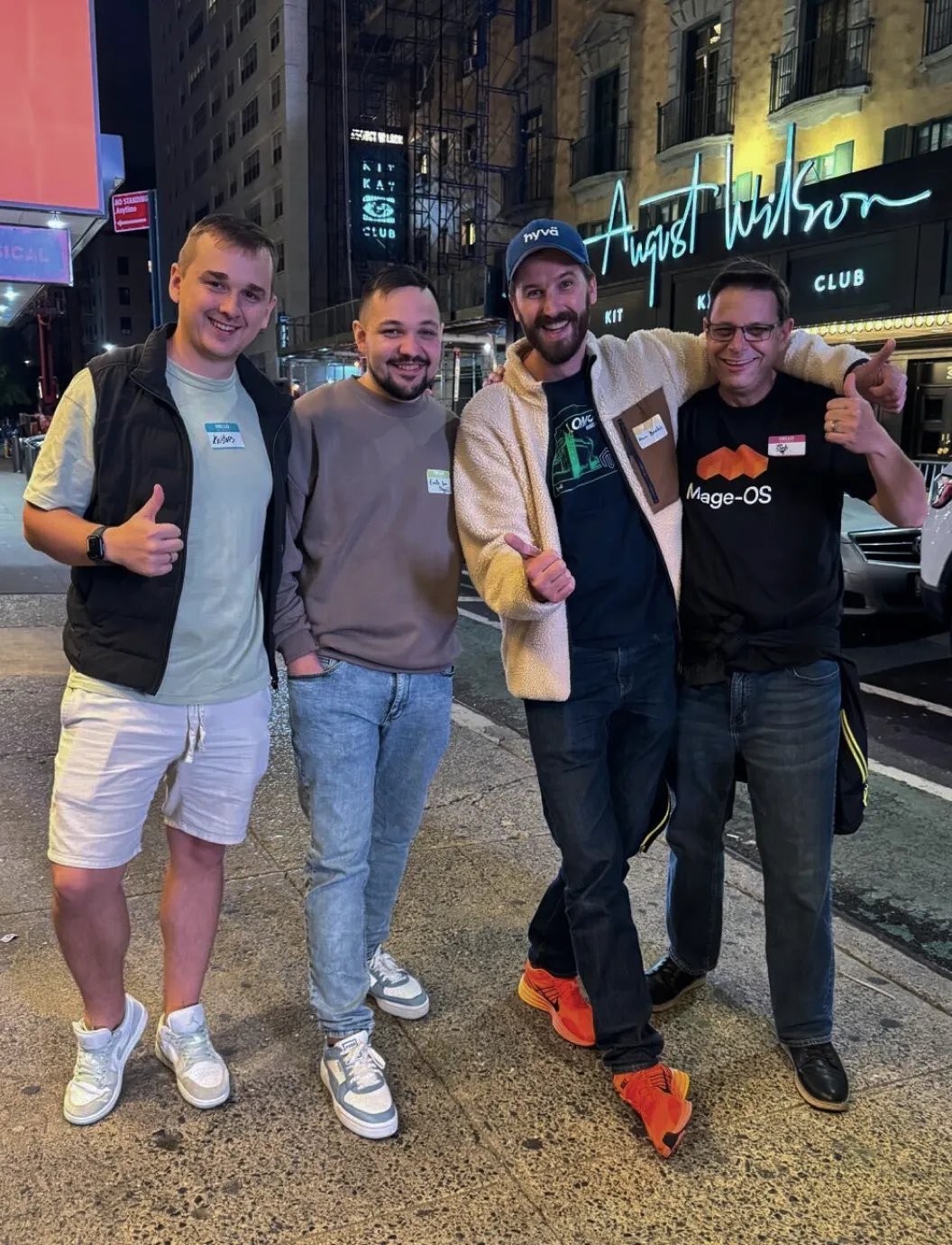 Four men standing outdoors at night, smiling and giving thumbs-up on a city sidewalk.