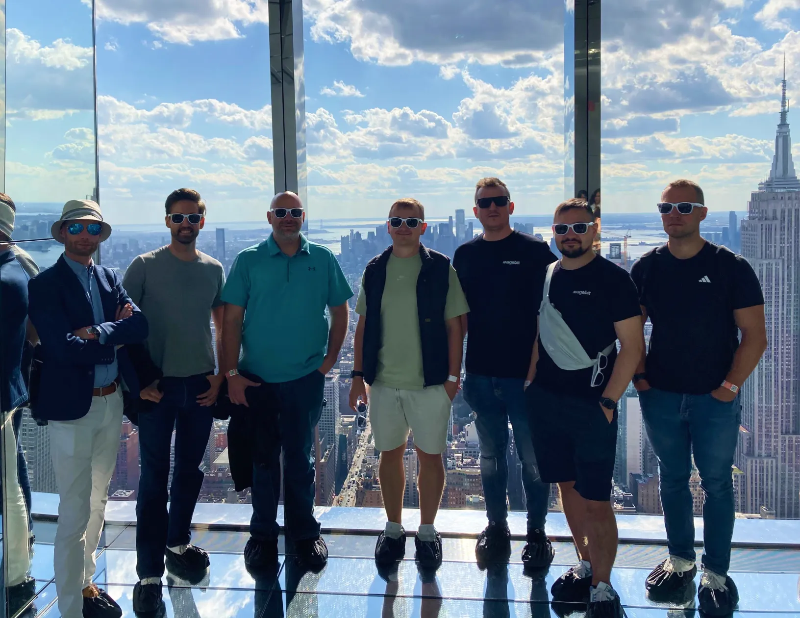 Group of seven men wearing sunglasses standing indoors with a city skyline view behind them.